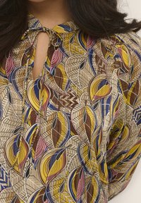 Blouse imprimée avec un col en forme de goutte d'eau. Les couleurs comprennent le jaune, le marron, le rose et le bleu avec des motifs de feuilles et un tissu texturé.