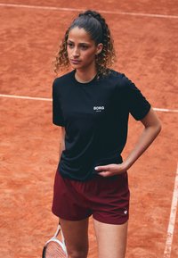 Svart idretts T-skjorte med hvitt logo, kombinert med maroon shorts. Modellen står på en leirtopps tennisbane, og holder en tennisracket.