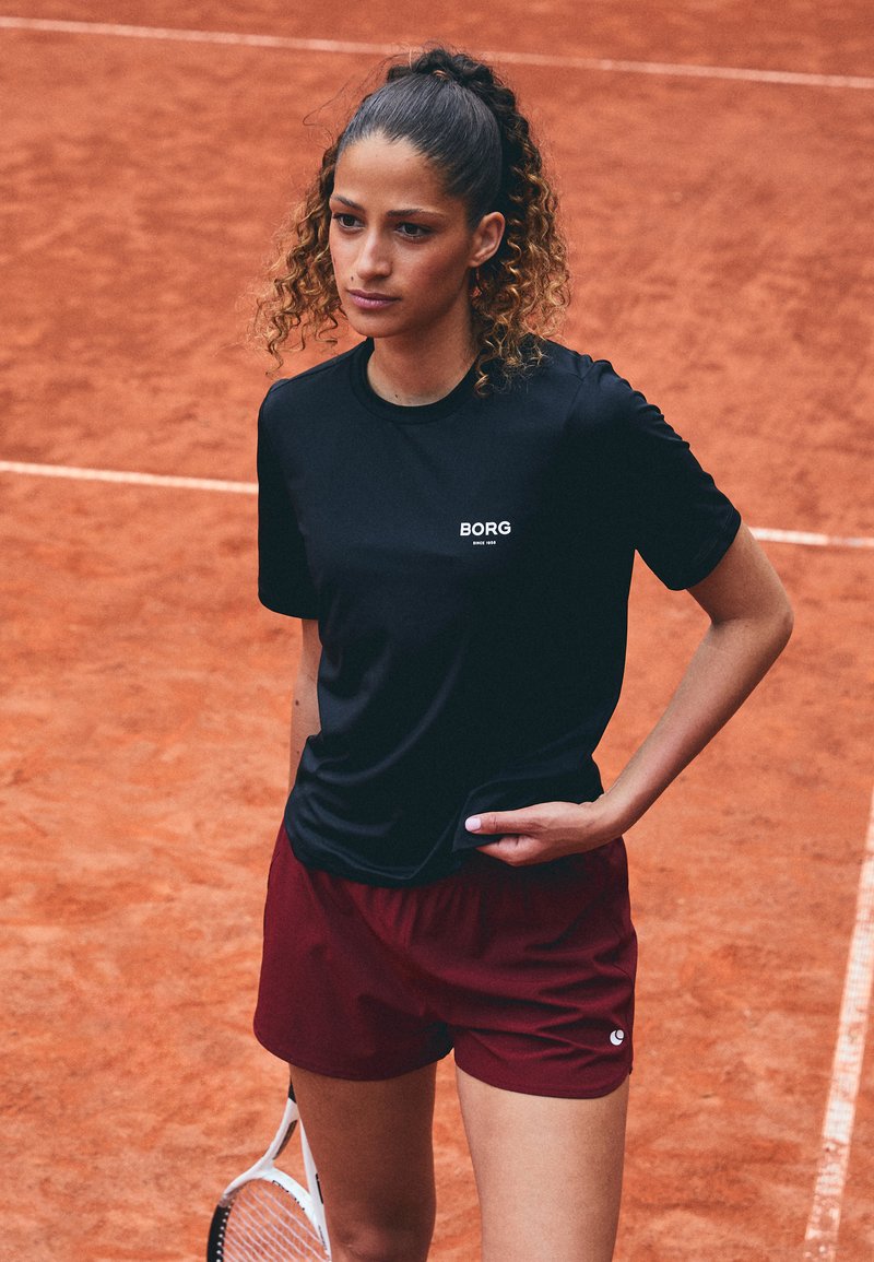Svart idretts T-skjorte med hvitt logo, kombinert med maroon shorts. Modellen står på en leirtopps tennisbane, og holder en tennisracket.