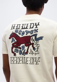 Άτομο που φοράει κρεμ πουκάμισο με μια κόκκινη απεικόνιση αλόγου και στιλιζαρισμένο κείμενο που λέει "Howdy Scotch & Soda Amsterdam Howdy" στην πλάτη.