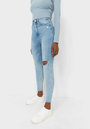 Personne portant un jean skinny bleu clair déchiré, des baskets compensées blanches et un haut bleu clair à manches longues sur un fond uni.