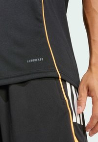 Černé sportovní tričko s oranžovým akcentovým švem a štítkem "AEROREADY". Detailní záběr ukazuje hladkou strukturu a střih, ruka odpočívající na černých šortkách ve stejném odstínu.