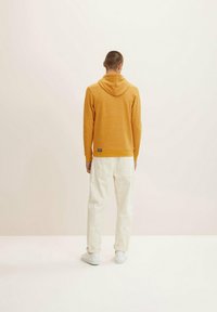 Sudadera amarilla con textura y cordones, con puños acanalados, combinada con pantalones anchos blancos y zapatillas blancas. Vista por la parte de atrás.