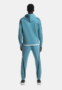 Homme portant un sweat à capuche bleu ciel et un pantalon assorti avec des rayures noires, vu de dos, se tenant contre un fond blanc.