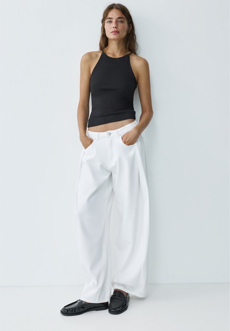 Femme debout contre un mur blanc, portant un haut noir sans manches, un pantalon blanc large, des mocassins noirs, les mains dans les poches, regardant droit devant.