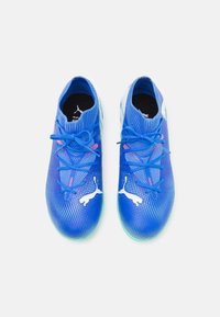 Zapatillas deportivas azules con una parte superior de malla texturizada, puntera redonda y cordones planos. Presenta un logo blanco y una suela iridiscente.