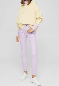 Světlé žluté mikiny s kapucí a šňůrkami, přes světlý fialový dlouhý rukáv, doplněné světlým fialovým úzkými džíny a bílými teniskami.