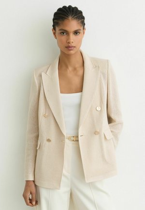 Femeie cu păr împletit poartă un sacou bej texturat, cu dublu rând de nasturi, un top alb cu dungi și pantaloni crema cu o curea asortată.