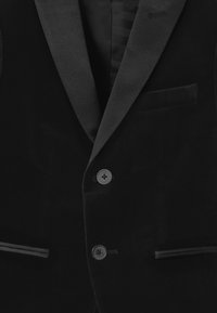 Černý sametový smokingový sako se saténovými klopami, dvouknoflíkovým zapínáním a jednou náprsní kapsou. Hladká textura s jemným leskem.