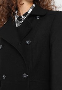 Svart dubbelknäppt kappa i ullliknande tyg, med stora svarta knappar och slagkrage. En rutig skjorta syns under.