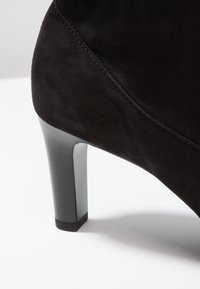 Scarpa nera in suede con tacco alto e texture liscia, caratterizzata da un design elegante e un tacco affusolato, parzialmente visibile nell'immagine.