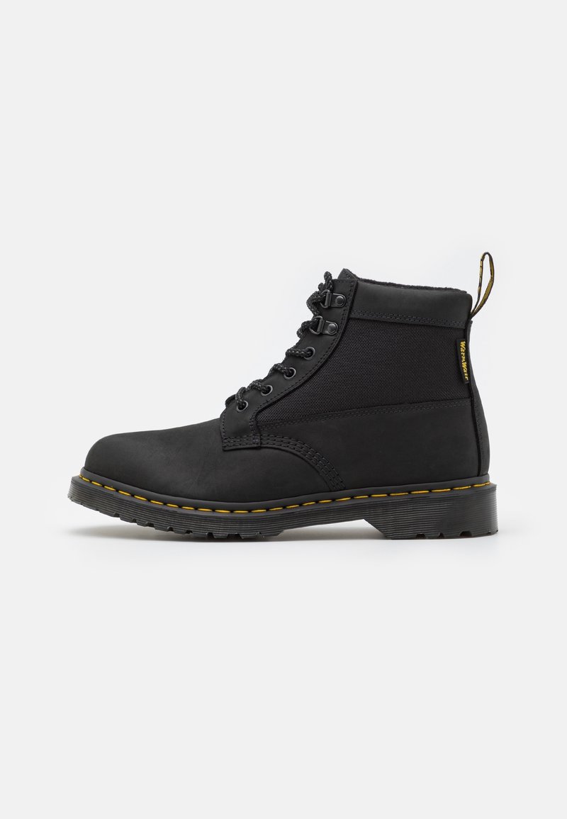 Bottine noire en cuir et tissu avec surpiqûres jaunes, lacets à l'avant et languette à l'arrière sur une semelle plate et robuste.