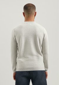 Hombre con cabello corto que lleva un suéter de manga larga texturizado en gris claro y pantalones oscuros, de espaldas contra un fondo simple.