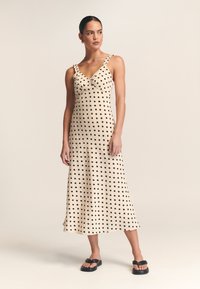 Beige jurk met polka dots, met een aansluitende lijfje en ruches op de schouderbanden, voorzien van een vloeiende zoom en gemaakt van lichtgewicht stof. Zwarte sandaaltjes gedragen.