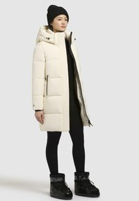 Vit vadderad jacka med huva, med dragkedja och sidofickor. Bärs över en svart outfit och matchas med svarta stövlar och en stickad mössa.