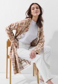 Lange, open voorkant cardigan in lichtbruin met geometrisch wit patroon. Gedragen over een wit T-shirt en gecombineerd met witte, op maat gemaakte broek.