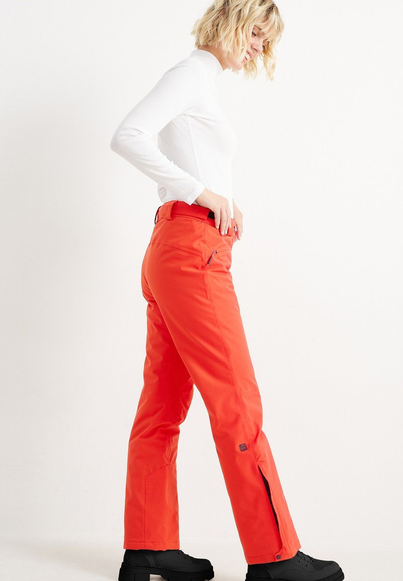 Helle orange Skihosen aus wasserdichtem Material, mit geradem Schnitt, seitlichen Reißverschlüsse und einem breiten Bund. Schwarze Schuhe runden den Look ab.