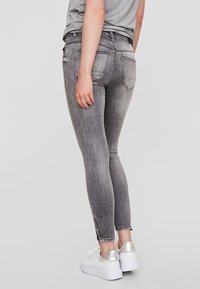 Jeans skinny de ganga cinza com um acabamento desbotado, apresentando bolsos traseiros e fechos laterais, combinados com ténis brancos e metálicos.