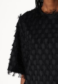Haut noir en tissu texturé avec un motif à pois et des bords effilochés. Manches courtes et col rond.