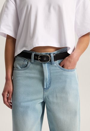 Černý kožený pásek se stříbrnou čtvercovou přezkou, nošený s bílým krátkým tričkem a světle modrými džínami s vysokým pasem.