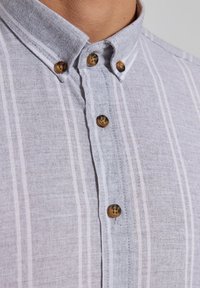 Gros plan sur une chemise gris clair boutonnée, avec des rayures blanches verticales et des boutons marron sur le col et la patte de boutonnage avant.