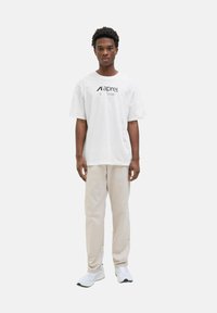 Weißes Baumwoll-T-Shirt mit bedrucktem Logo und Text, kombiniert mit hellbeigen Hosen und weißen Sneakers, präsentiert von einem stehenden Modell.