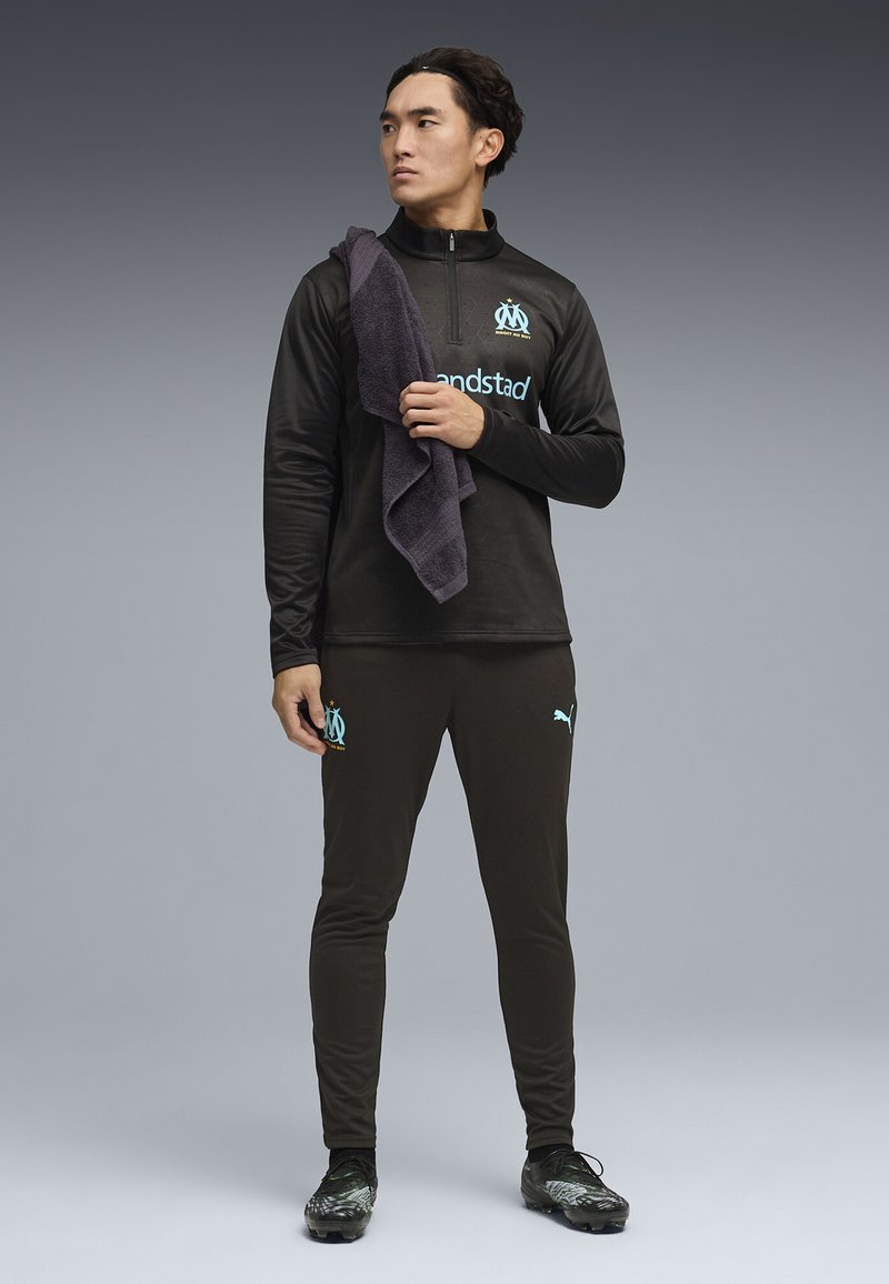 Atlet bărbat în echipament sportiv negru cu sigla „Marseille” și brandul Puma, ținând un prosop închis la culoare pe umăr, privind în lateral.