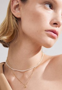 Colliers en or et en perles superposés, avec un pendentif en forme de cœur sur la chaîne inférieure. Peau lisse et cheveux clairs visibles.