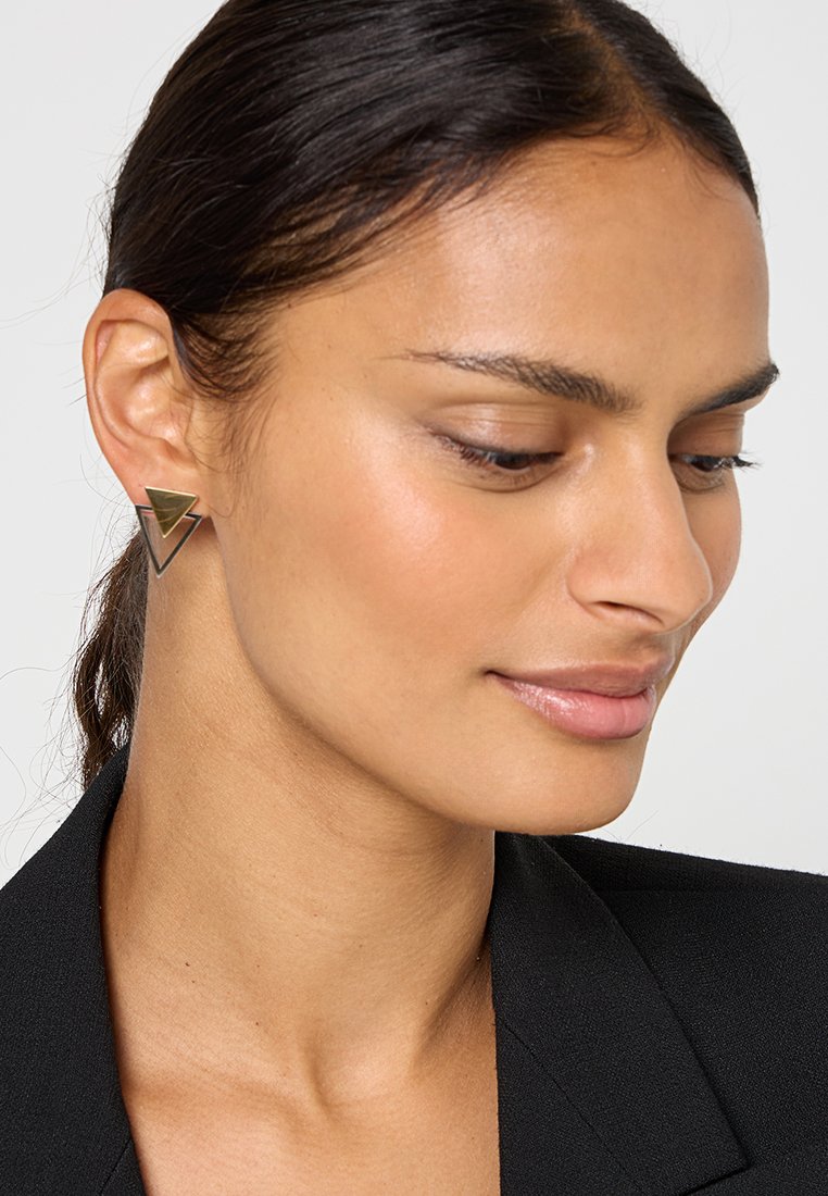 Geometrische Metallohrringe mit einem dreieckigen Design in gebürstetem Goldton, am linken Ohr getragen vor einem dunklen Blazer-Hintergrund.