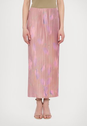 Kvinde iført en lyserød plisseret midi-nederdel med lilla blomsterprint, beige ankelsandaler med rem og en beige top, stående mod en hvid baggrund.