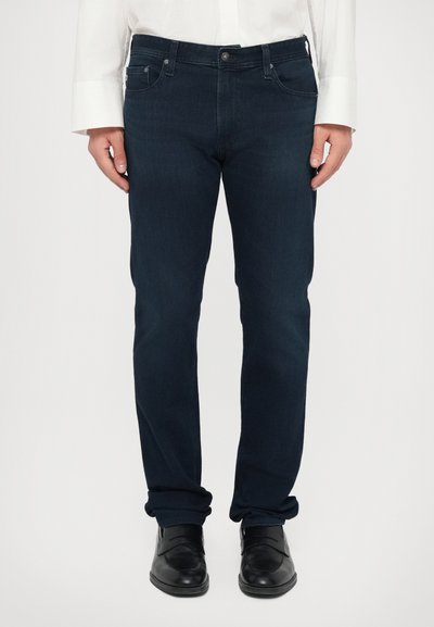 Jeans in denim blu scuro con taglio dritto, caratterizzati da un design a cinque tasche e dettagli di cucitura sottili. Abbinati a scarpe nere e una camicia bianca.