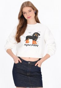 Sudadera blanca corta con un gráfico de un perro negro con botas naranjas, con anotaciones de texto sobre sus características. Falda de mezclilla oscura.