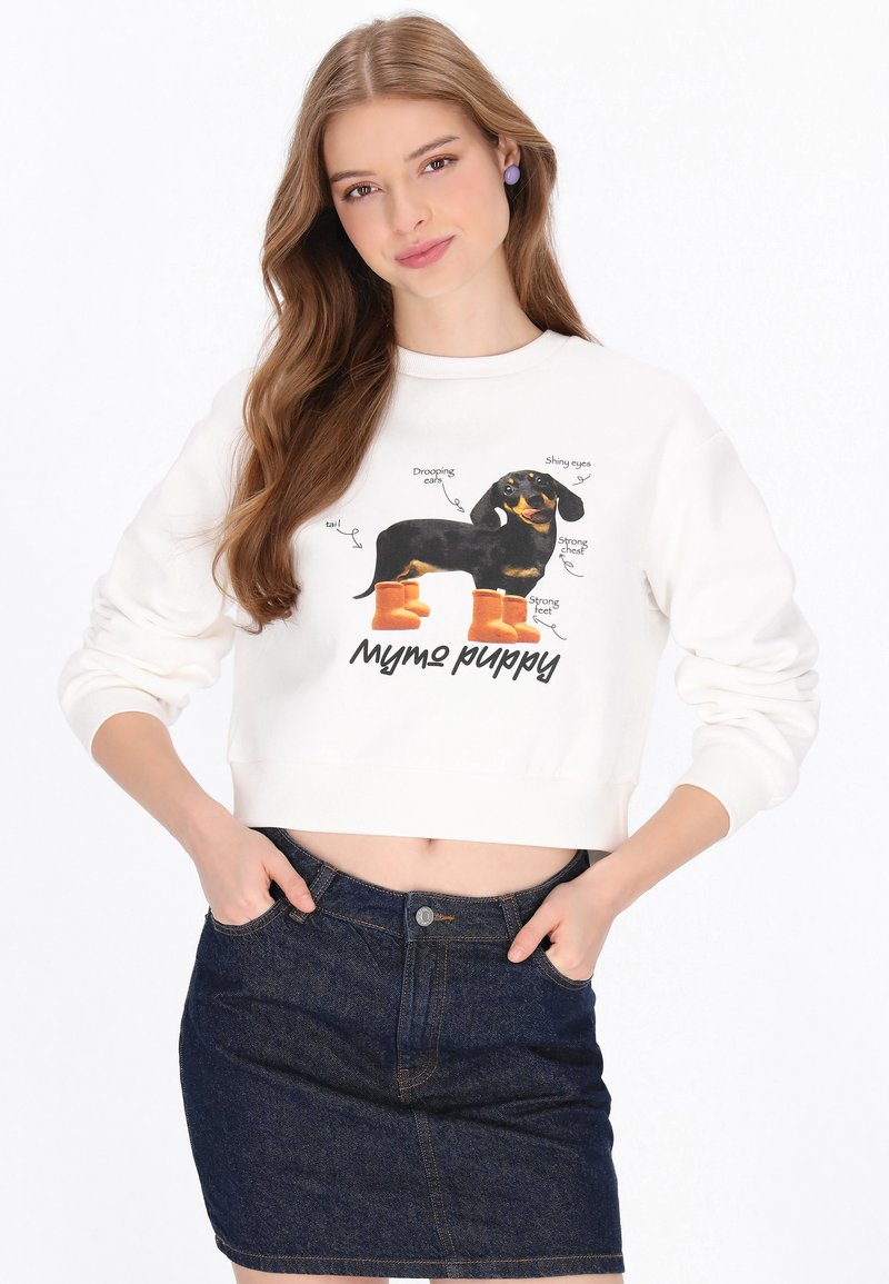 Sudadera blanca corta con un gráfico de un perro negro con botas naranjas, con anotaciones de texto sobre sus características. Falda de mezclilla oscura.