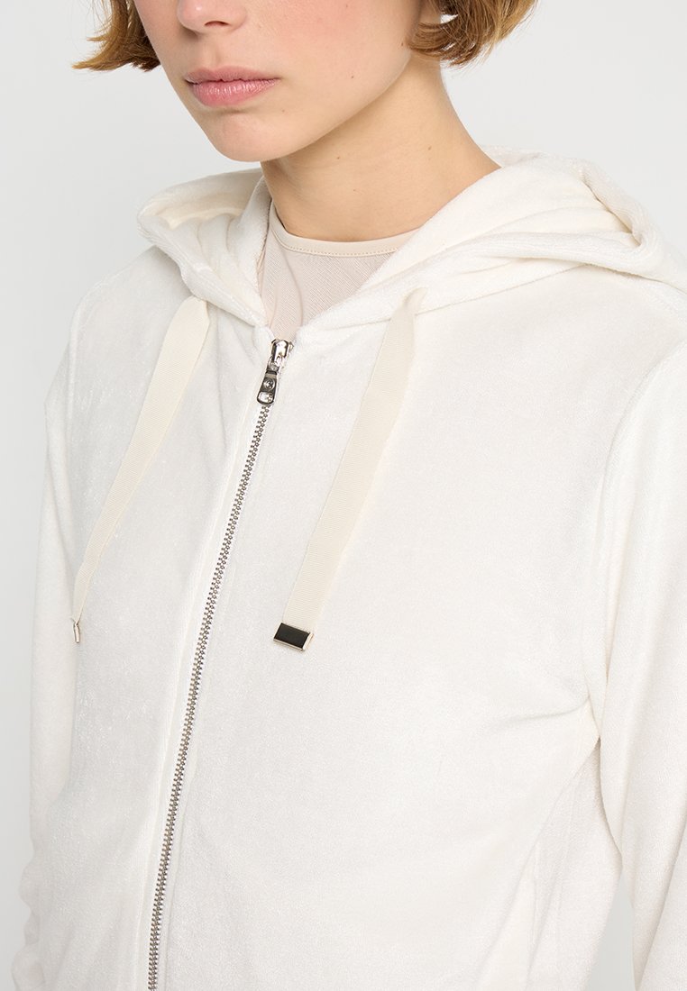 Felpa in pile crema con zip, cordini e dettagli argento. Presenta una texture morbida e una vestibilità rilassata.