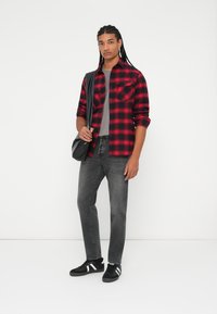 Rød og svart rutete flanellskjorte, grå t-skjorte, svarte jeans, svarte sko med hvite striper. Modell holder en svart skulderveske.
