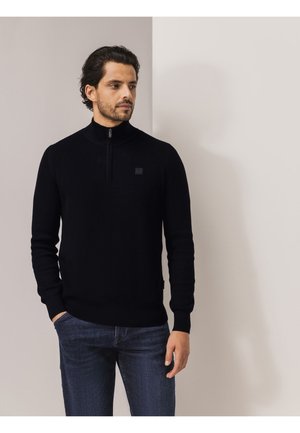 Schwarzer Strickpullover mit hohem Kragen und halbnahtigem Reißverschluss-Design. Verfügt über ein strukturiertes Muster und ein kleines Logo auf der Brust. Getragen zu blauen Jeans.