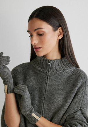Grau gestrickte Handschuhe mit gerippten Bündchen, getragen von einer Person mit einem passenden Rollkragenpullover, sind mit einem kleinen Etikett mit dem "FALCONERI"-Logo versehen.