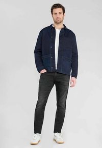 Marineblauwe katoenen jas met knoopsluiting, twee voorzakken en een ontspannen pasvorm, gecombineerd met zwarte jeans en witte sneakers.