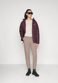 Maroon puffer jacket with a hood, beige textured top, beige pants, and black sneakers. Model wears oversized black sunglasses.