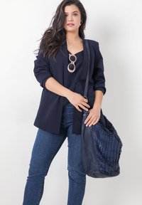 Marineblazer met opgerolde mouwen, gecombineerd met een zwarte top en donkere jeans. Houdt een gestructureerde marineblauwe tas met een geweven ontwerp en een hengsel.