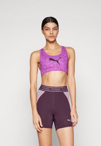 Sujetador deportivo morado con logo negro, combinado con pantalones cortos oscuros de cintura alta con acentos morados y rosas y costuras en contraste.