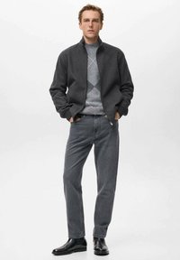 Giacca grigia in misto lana con colletto rialzato, indossata sopra un maglione leggero grigio con motivo argyle, abbinata a jeans grigio scuro e stivali neri.