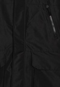 Casaco impermeável preto com uma textura suave, apresentando um fecho frontal, dois bolsos grandes e um bolso lateral com fecho.