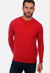 Pull rouge à manches longues avec col rond, fabriqué dans un matériau doux, présentant une texture lisse et une coupe ajustée. Associé à un jean foncé.