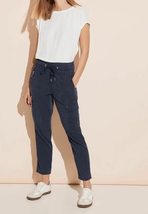 Morskie spodnie cargo z elastycznym pasem i sznurkiem do regulacji. Biała bluzka z krótkim rękawem. Model nosi białe sneakersy z brązowymi akcentami.