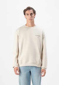 Sweat-shirt beige à col rond et manches longues, avec un petit logo noir sur la poitrine. Porté avec un jean bleu clair.