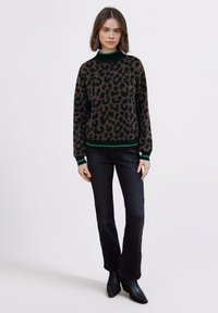 Brązowy sweter w cętki lamparta z czarnym, prążkowanym dekoltem i zielonymi akcentami, zestawiony z ciemnymi, dzwonowatymi dżinsami oraz czarnymi, spiczastymi botkami.