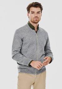 Szary dzianinowy kardigan z zamkiem z przodu, ze ściągaczami przy mankietach i w pasie. Noszony na ciemnozielonej koszuli z kołnierzykiem, o miękkiej fakturze.