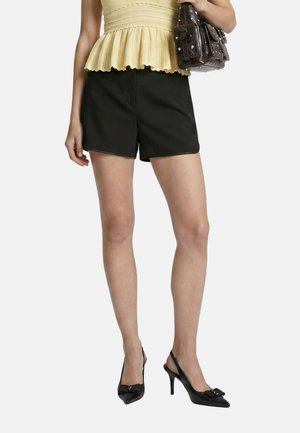 Donna con pantaloncini neri a vita alta, top giallo senza maniche in stile peplum, tacchi neri a punta e che tiene una borsa a mano marrone scuro.