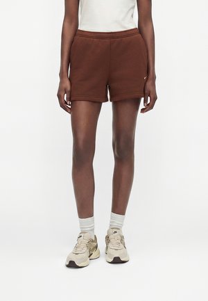 Person iført brune sportsshorts, hvide sokker, beige Nike-sneakers og en hvid top, stående mod en ensfarvet lys baggrund.